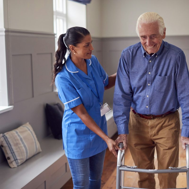 Senior Man At Home Using Walking Frame Being Helped By Female Care Worker In Uniform